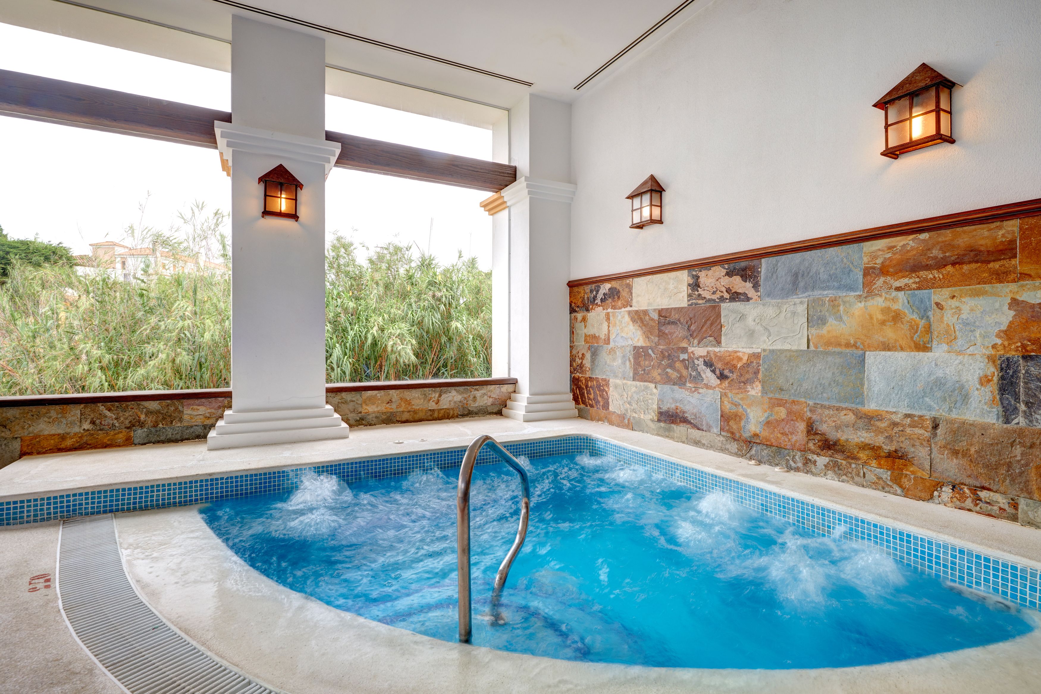 A hot tub with a stone wall and columns, a view of greenery outside, and two wall-mounted lanterns.