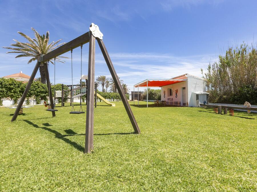 Un parque infantil con columpios y un tobogán, rodeado de césped y árboles.