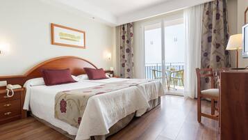 A hotel room with a large bed, a desk, a chair, and a balcony with curtains.