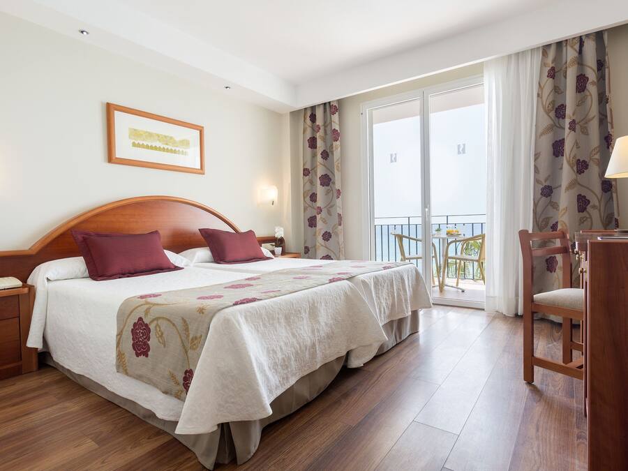 A hotel room with a large bed, a desk, a chair, and a balcony with curtains.