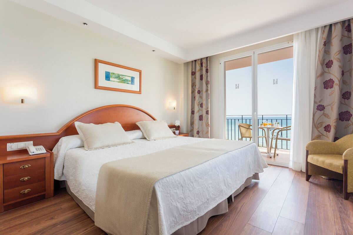 Een hotelkamer met een groot bed, een nachtkastje met telefoon, een stoel, een balkon met tafel en stoelen, en een schilderij aan de muur.