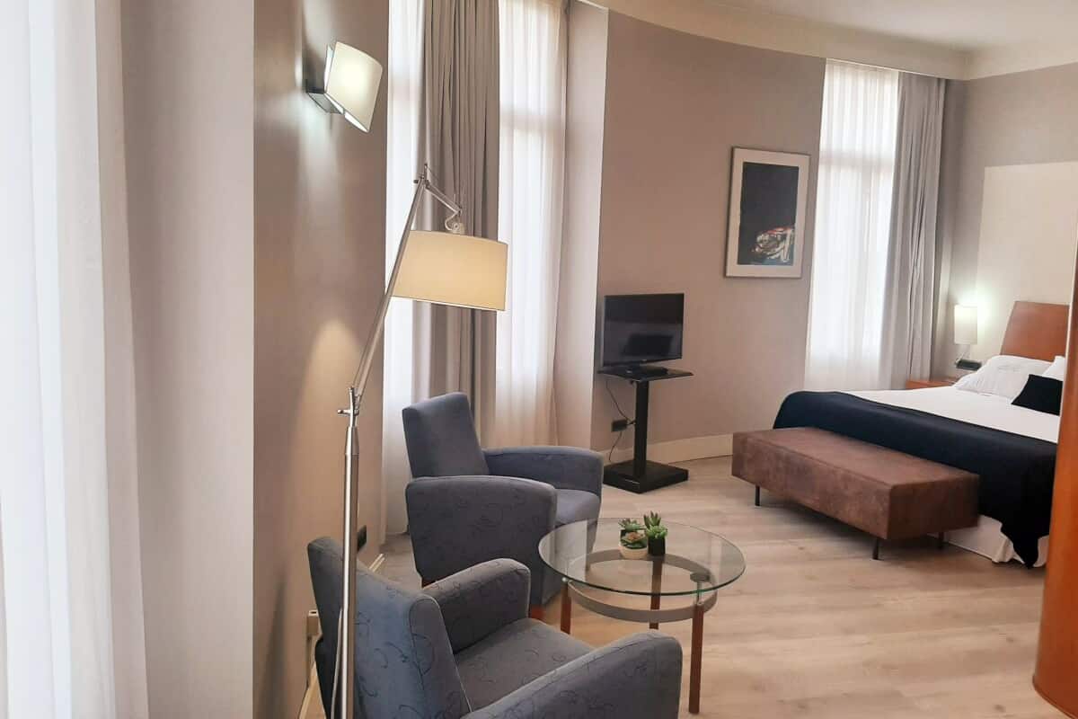 A hotel room with a bed, a desk, a chair, a sofa, a TV, and a lamp.