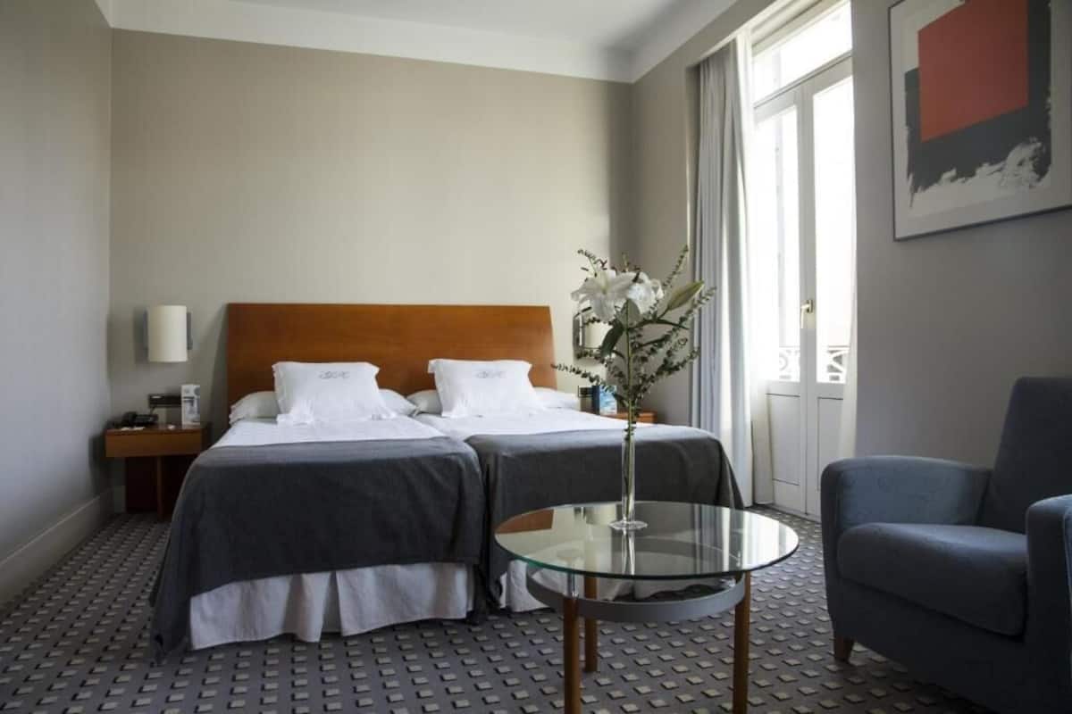 A hotel room with two beds, a glass table, a chair, a nightstand, a lamp, and a vase with flowers.
