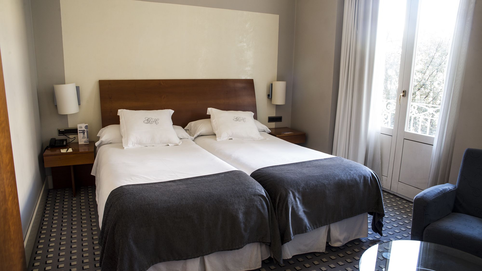 Habitación de hotel con dos camas, cabecera de madera, ropa de cama blanca, un escritorio con teléfono y lámpara, y una ventana con cortinas.