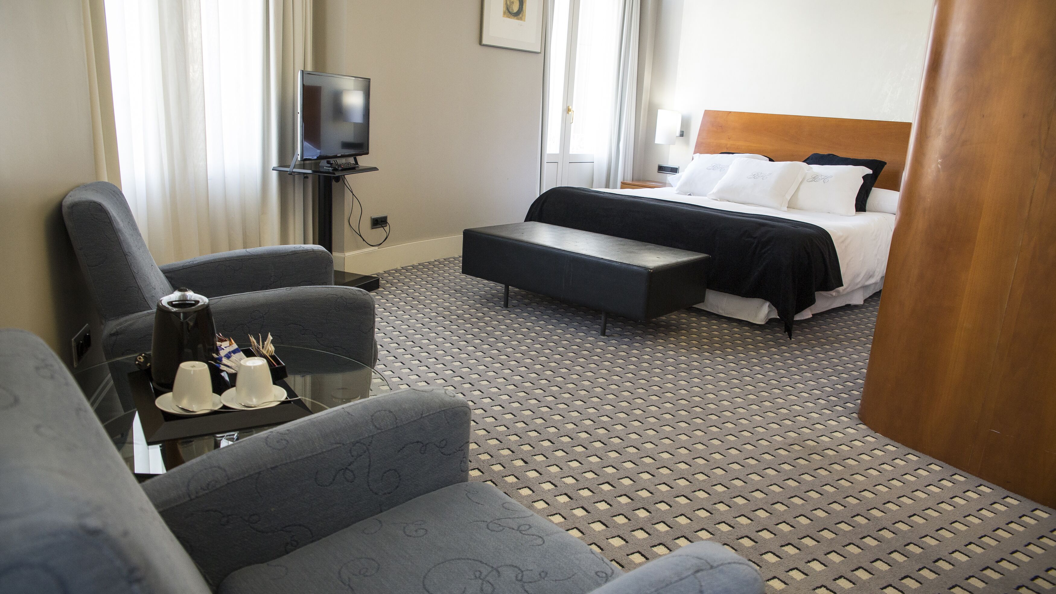 A hotel room with a bed, a TV, a desk with a chair, and a small table with a cup and saucer.