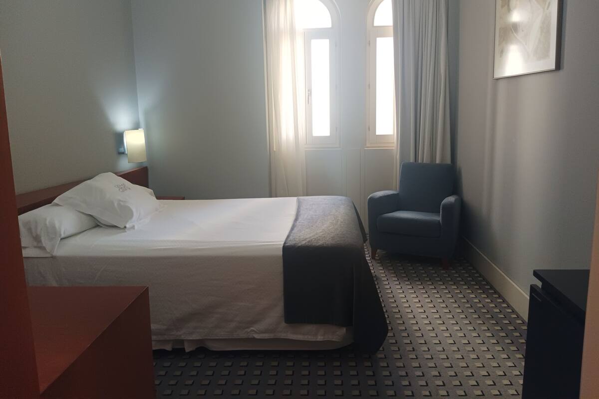 A hotel room with a bed, a chair, a desk, and a window with curtains.