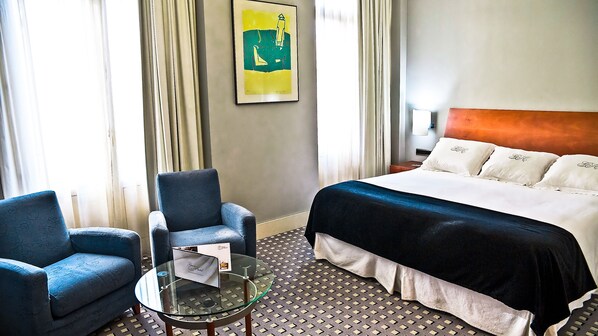 A hotel room with a bed, two armchairs, a glass table, and a painting on the wall.