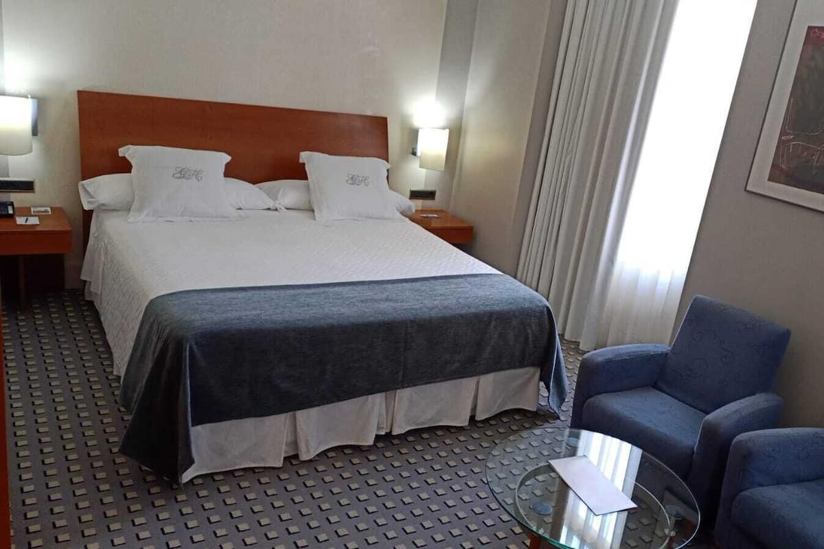 A hotel room with a large bed, two bedside tables, a desk with a chair, and a window with curtains.