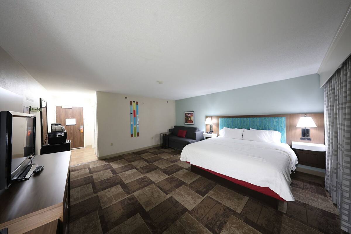 A hotel room with a bed, a desk with a TV, a chair, a nightstand, and a bathroom.