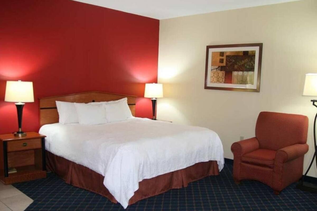 Una habitación de hotel con una cama grande, dos mesitas de noche con lámparas, un sillón rojo y un cuadro enmarcado en la pared.