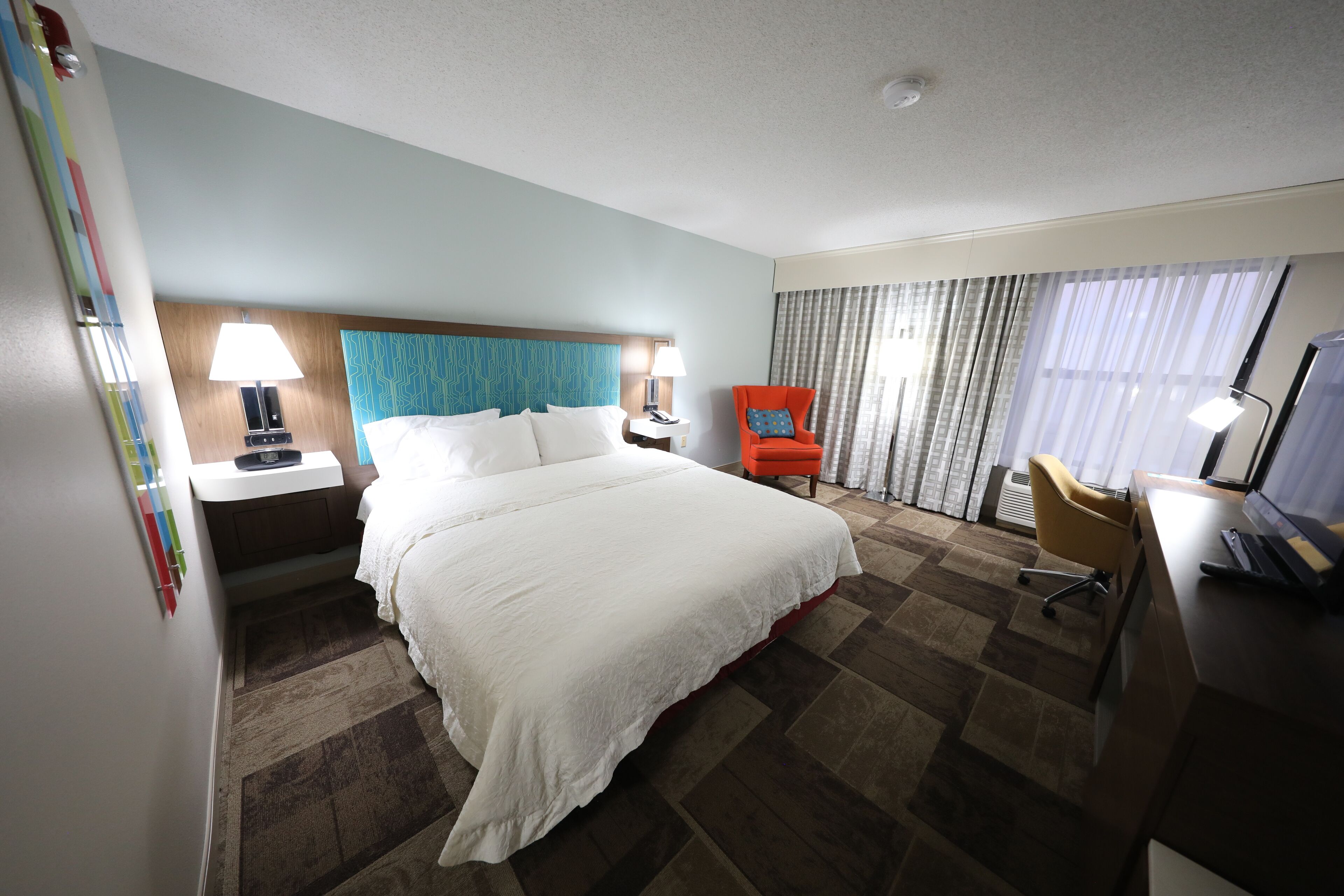 A hotel room with a large bed, a desk with a computer, a chair, a nightstand with a lamp, and a window with curtains.