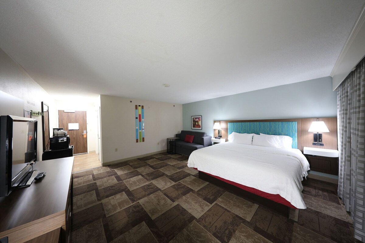 A hotel room with a bed, a desk with a TV, a chair, a nightstand, and a bathroom.