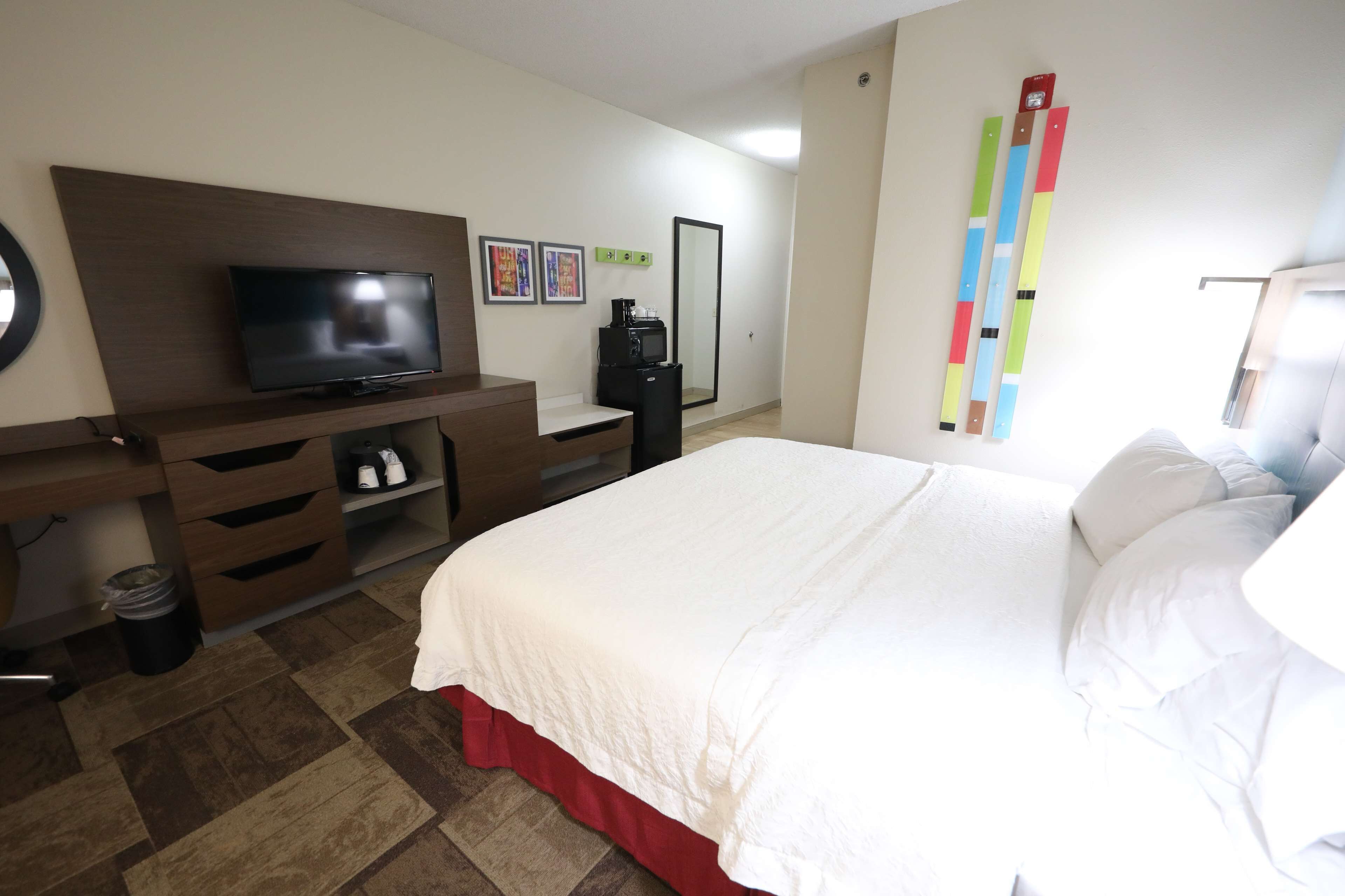 A hotel room with a bed, a TV, a desk with drawers, a trash bin, and a mirror.