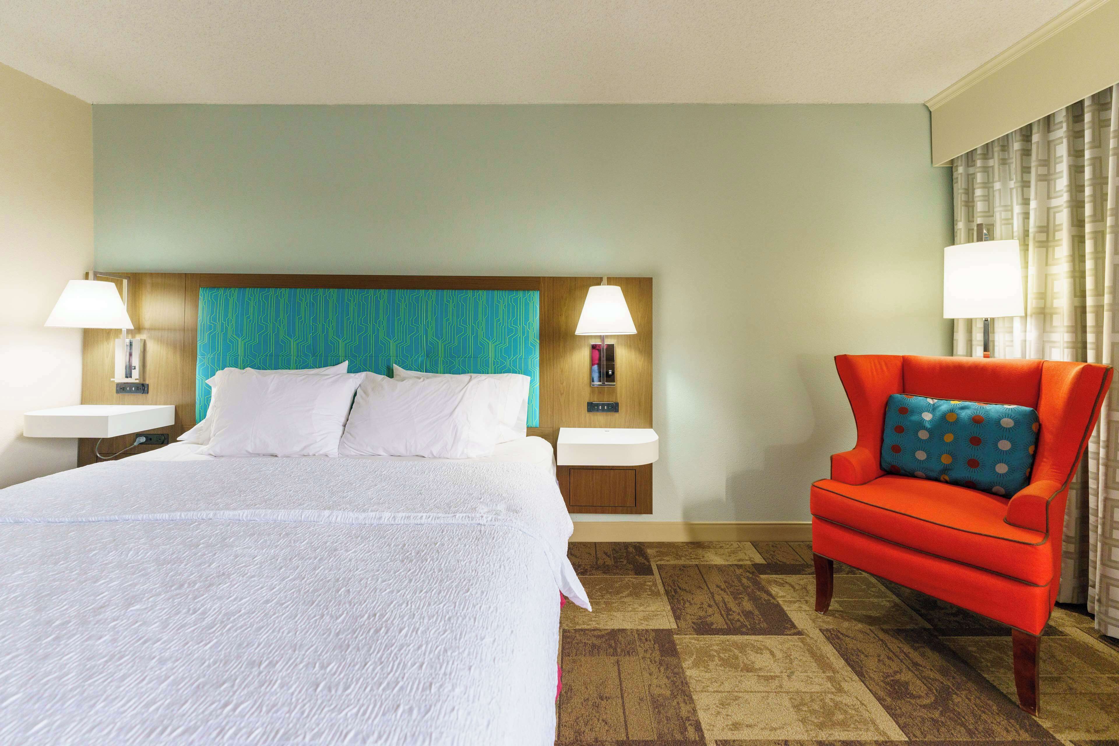A hotel room with a large bed, two bedside tables with lamps, a red armchair, and a patterned headboard.