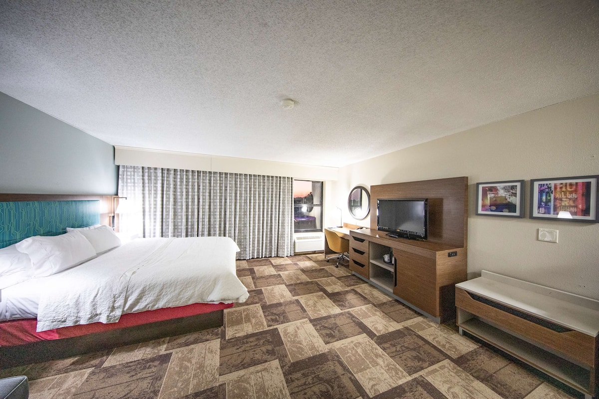 Una habitación de hotel con cama, escritorio, televisor y ventana con cortinas.