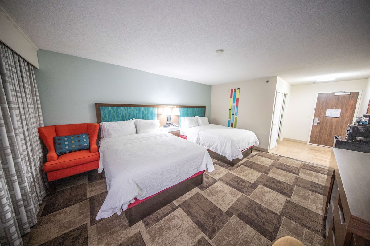 Habitación de hotel con dos camas, un sillón rojo, un escritorio y la puerta del baño.