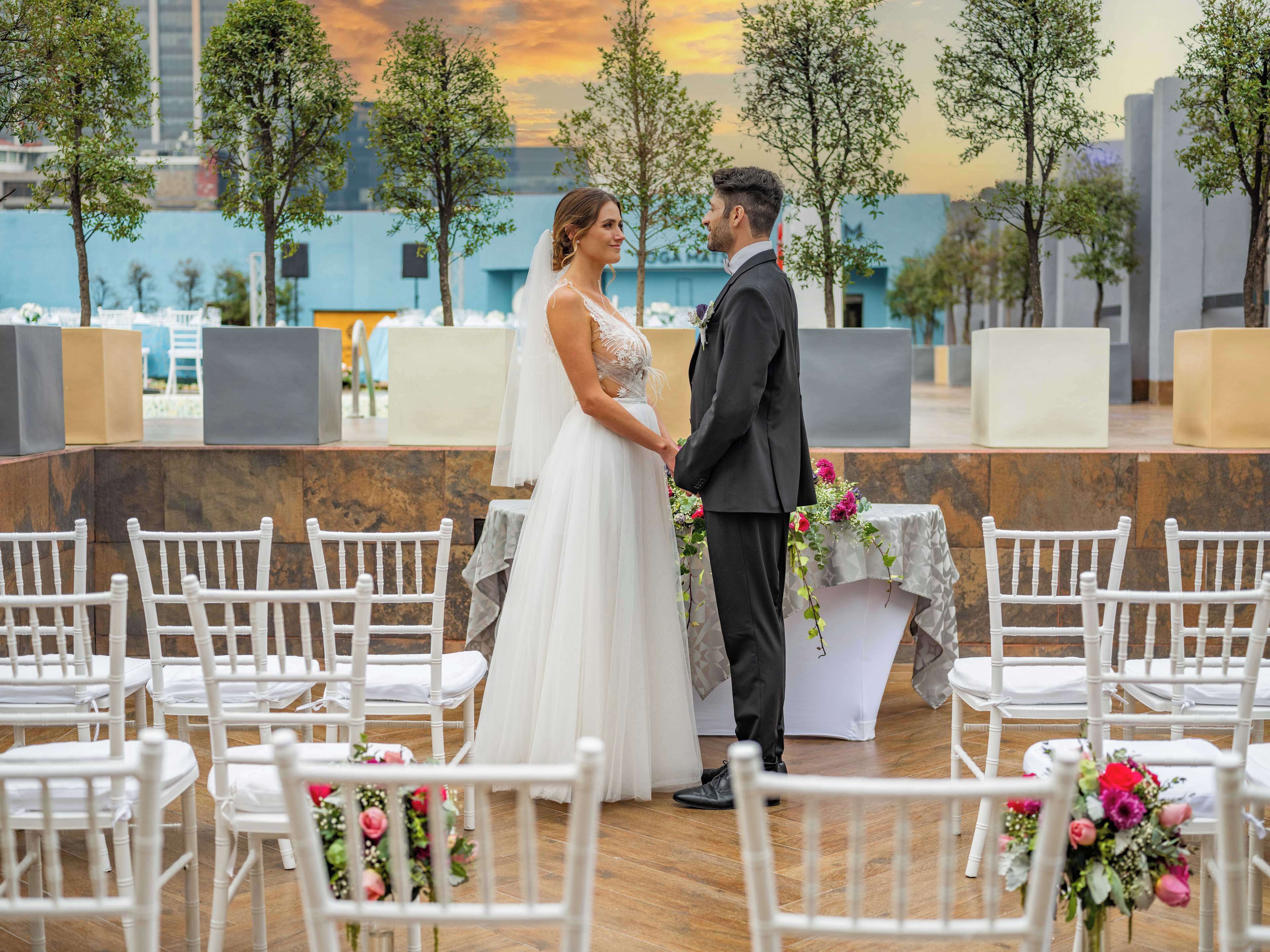 Uma cerimônia de casamento com um casal no centro, rodeado por cadeiras brancas e arranjos florais.