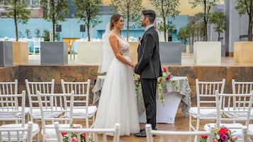 Uma cerimônia de casamento com um casal no centro, rodeado por cadeiras brancas e arranjos florais.