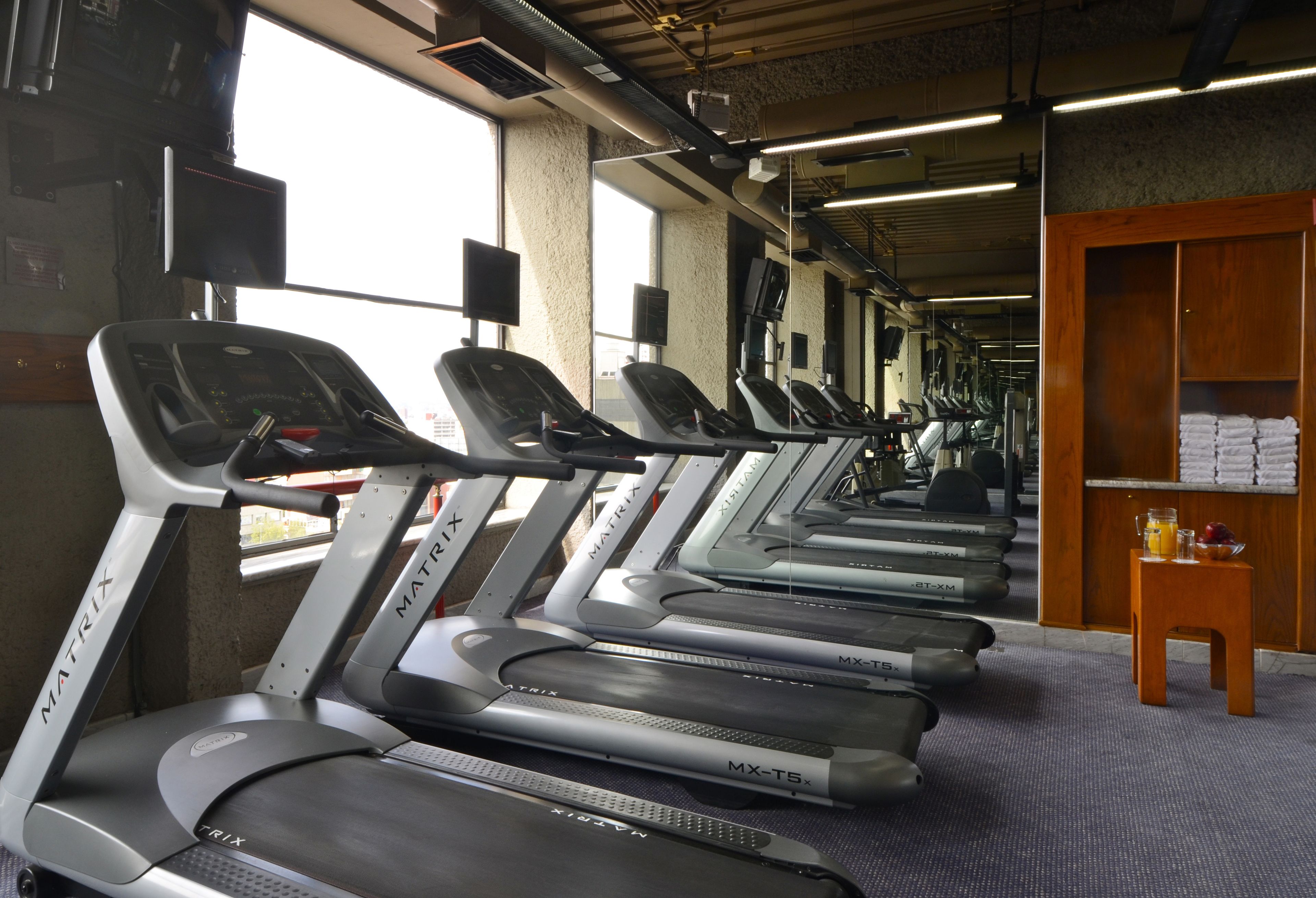 Varias caminadoras en un gimnasio, con una pequeña mesa y toallas a la derecha.