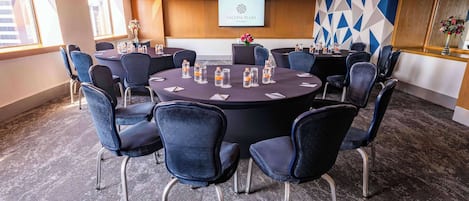 Un salón de conferencias con mesas redondas, sillas y una pantalla de proyector que muestra