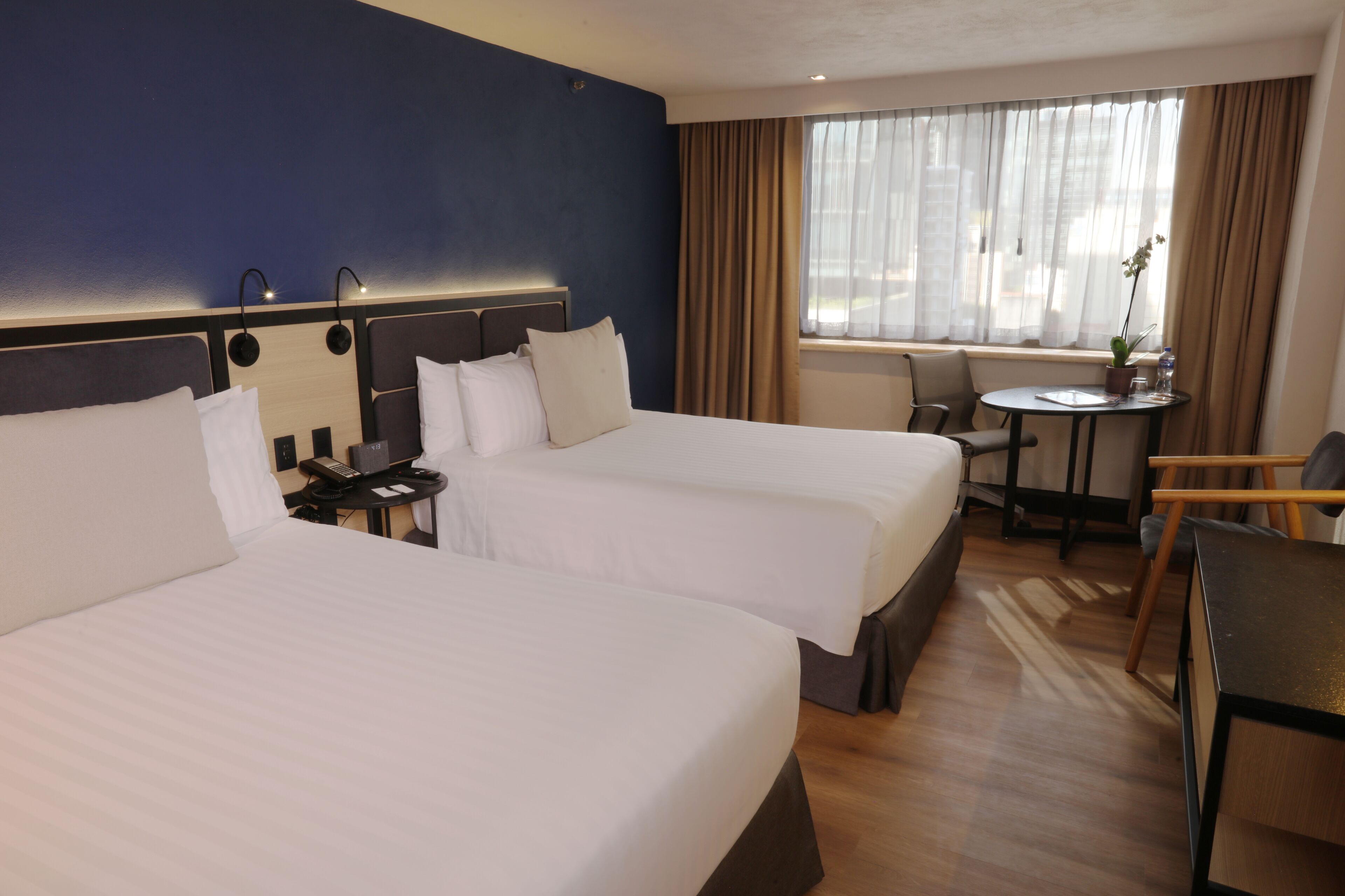 A hotel room with two beds, a desk, a chair, a small table, and a window with curtains.