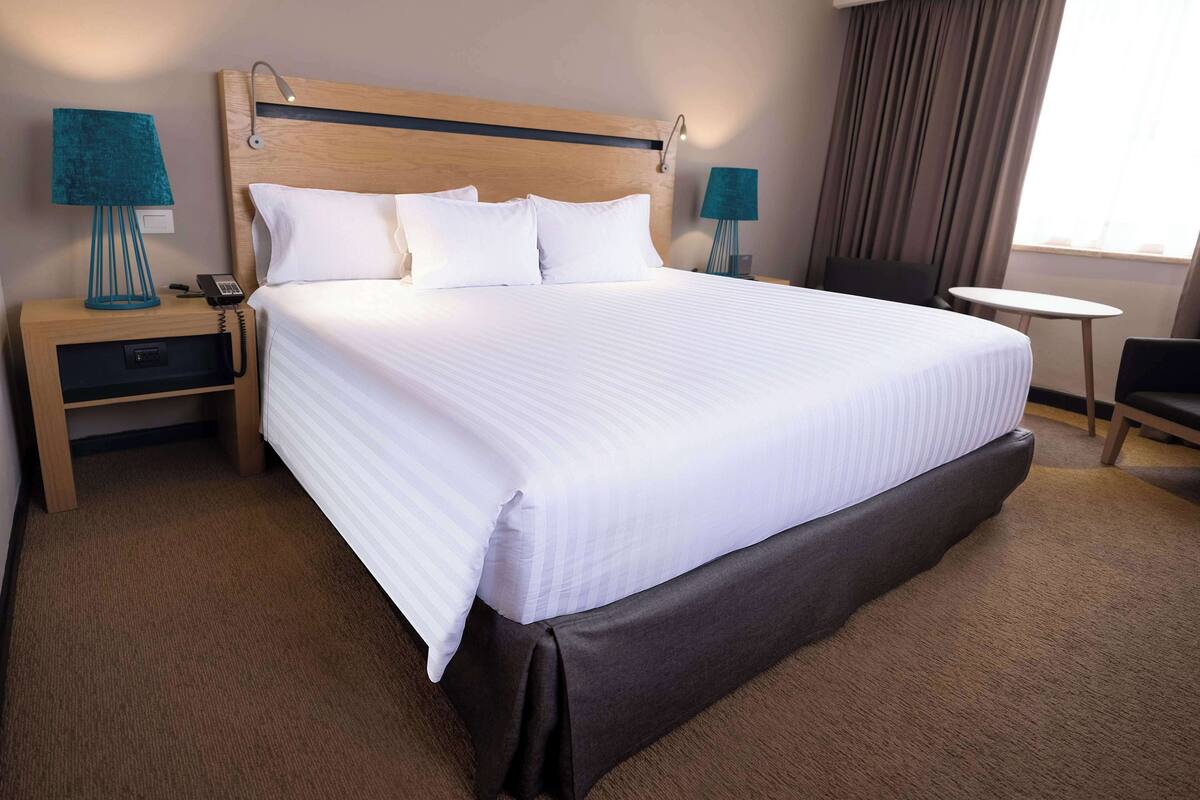 A hotel room with a large bed, two bedside tables with lamps, a small round table, a chair, and a window with curtains.