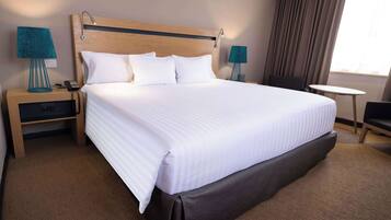 Quarto de hotel com uma cama grande, duas mesas de cabeceira com abajures, uma mesa redonda pequena, uma cadeira e uma janela com cortinas.