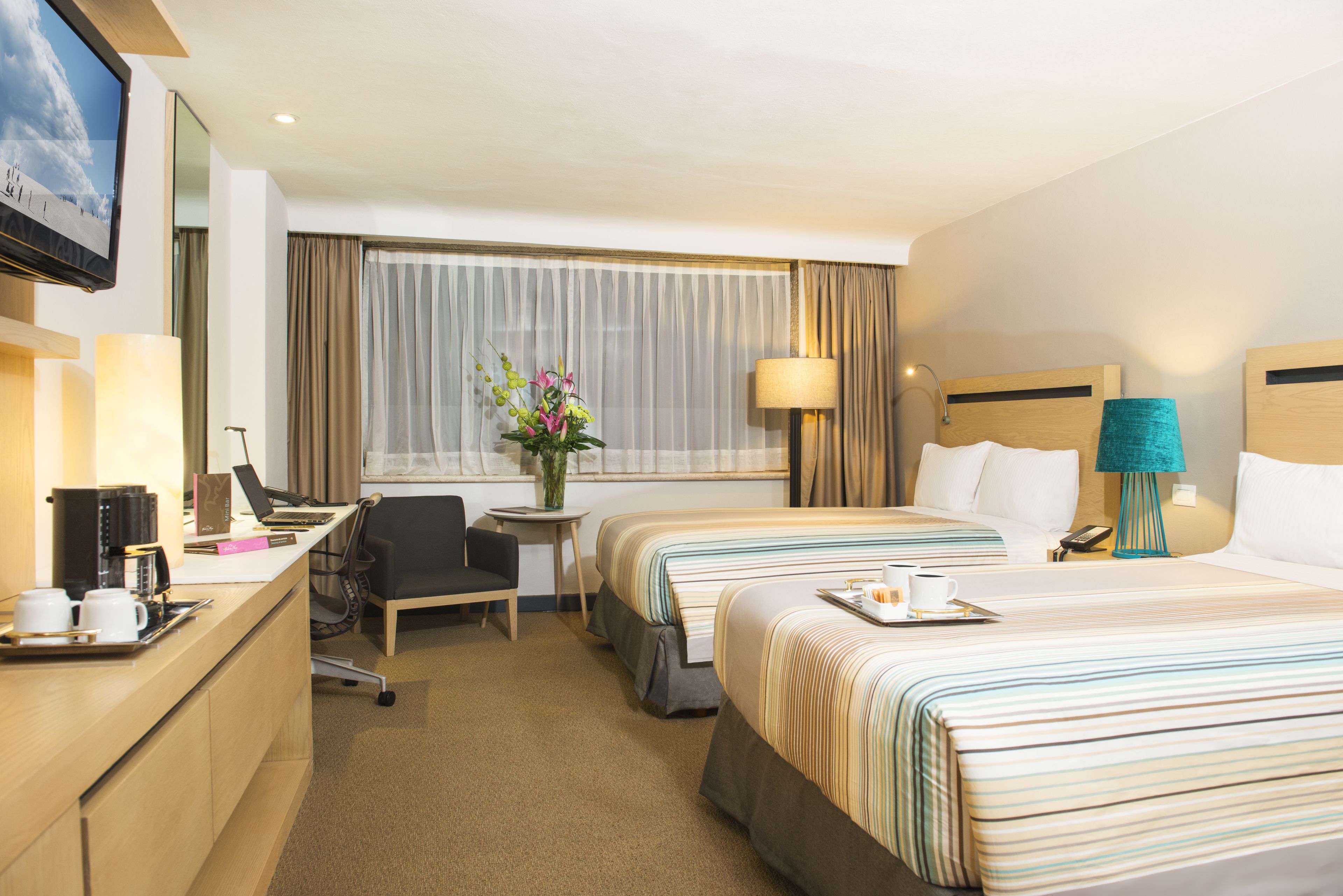 A hotel room with two beds, a desk with a laptop, a chair, a coffee maker, a vase with flowers, a lamp, and a TV on the wall.