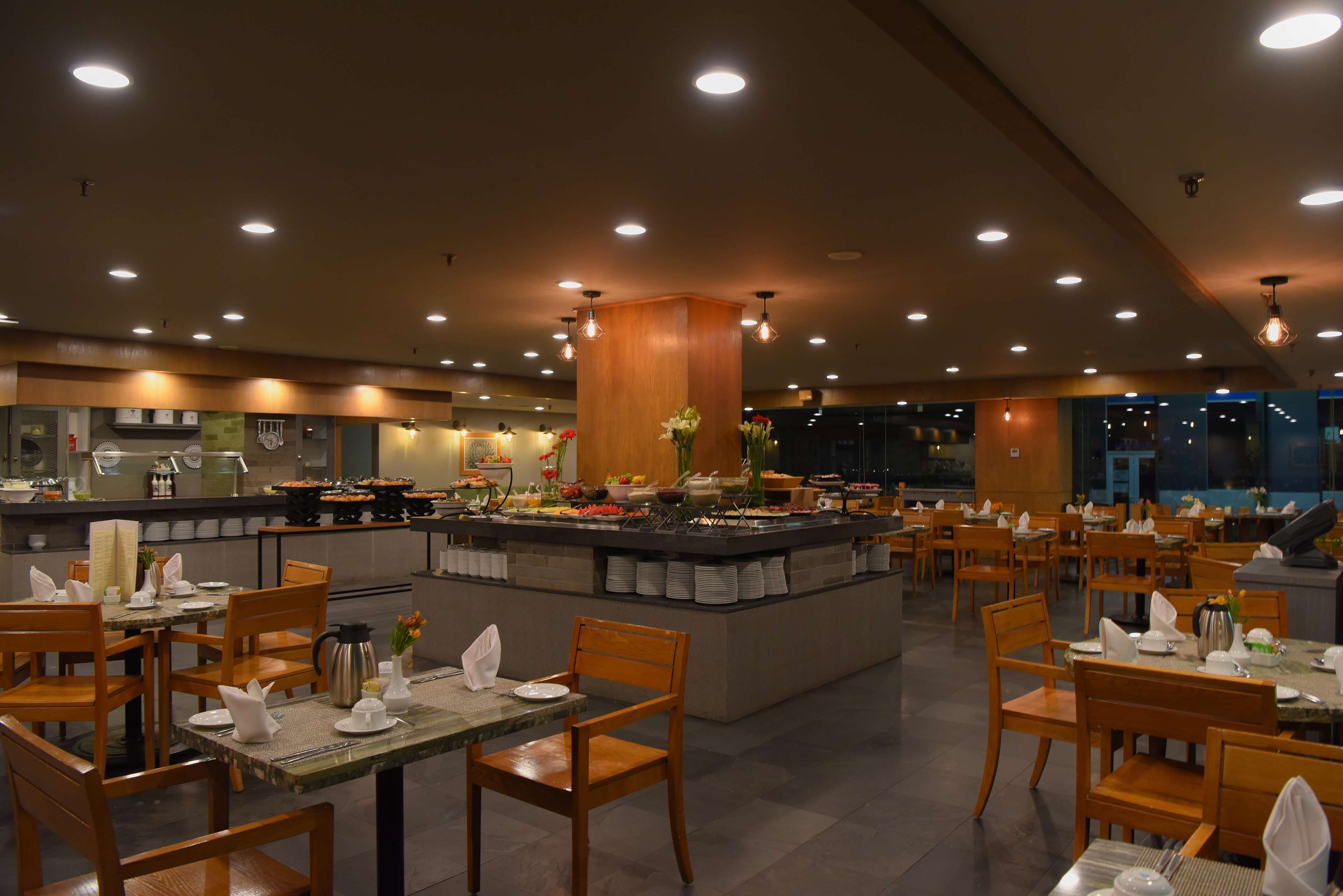 A restaurant with multiple dining tables and chairs, a central buffet area with various food items, and a kitchen in the background.