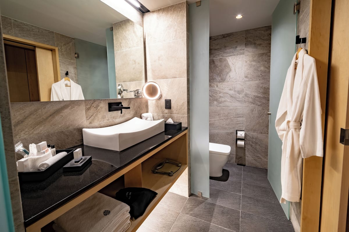 Ein modernes Badezimmer mit einem gebogenen Waschbecken, einem Waschtisch, einem Spiegel mit Beleuchtung, einer Toilette und einem Bademantel, der an der Tür hängt.