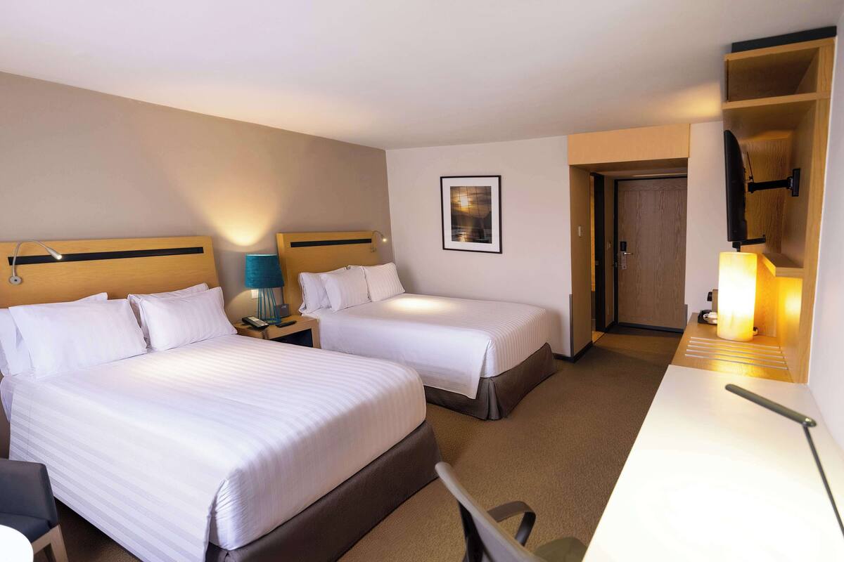 A hotel room with two beds, a desk, a chair, a lamp, and a framed picture on the wall.