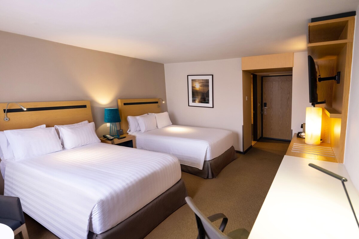 A hotel room with two beds, a desk, a chair, a lamp, a framed picture on the wall, and a door leading to another room.