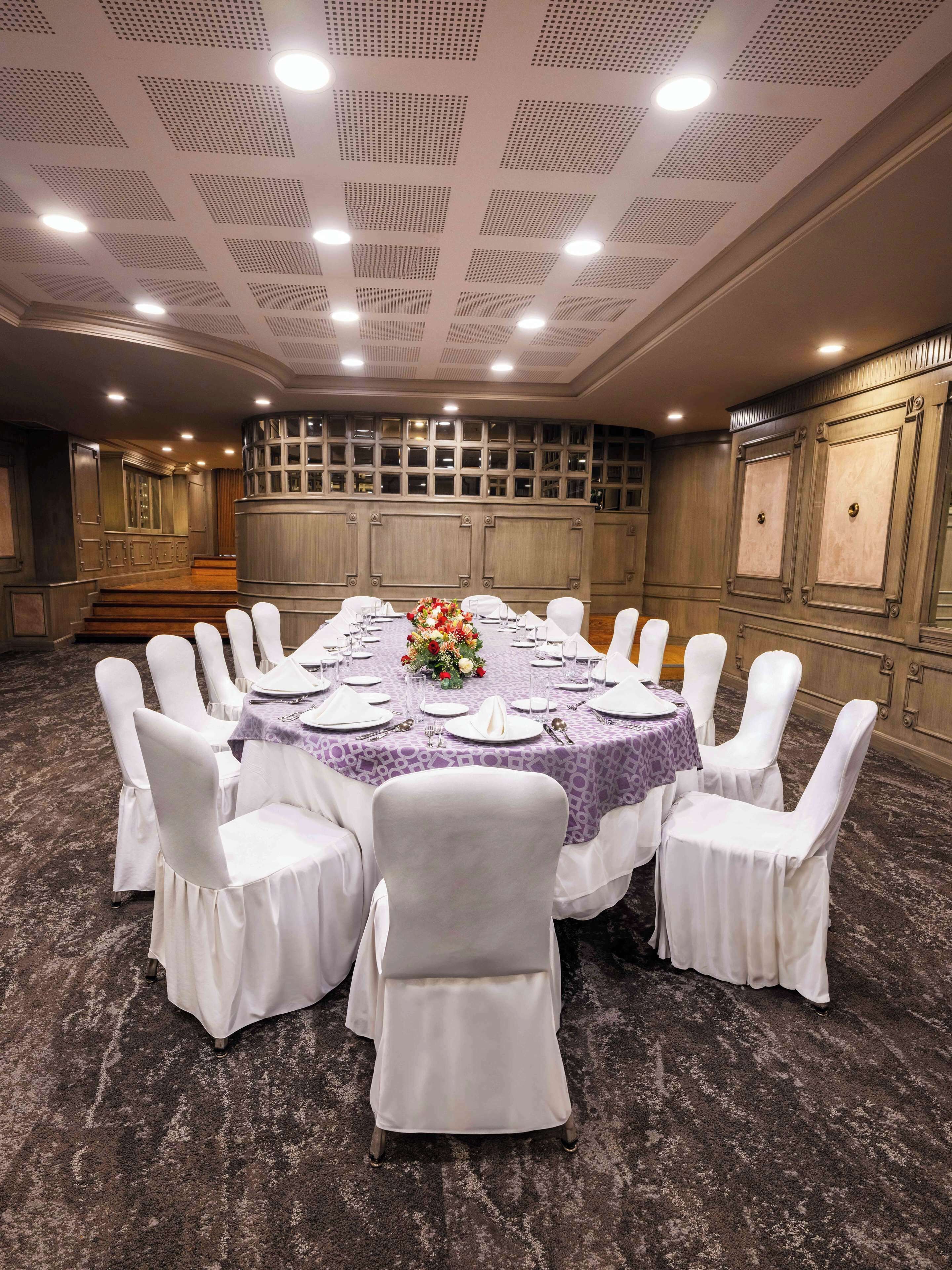 Un salón de conferencias con una mesa redonda, sillas blancas y un mantel morado.