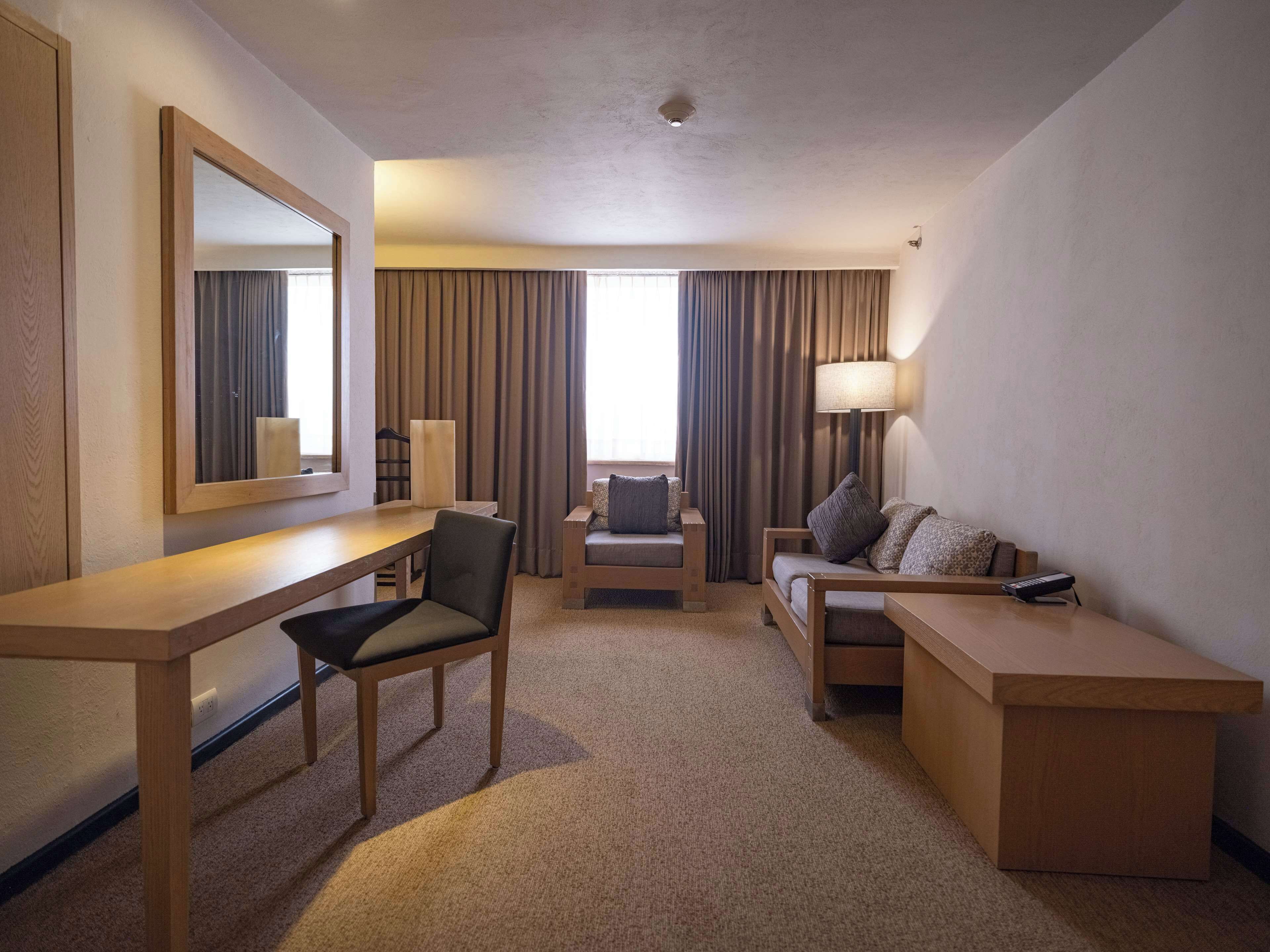 Quarto de hotel com escrivaninha de madeira, uma cadeira, um sofá, abajur e um espelho grande.