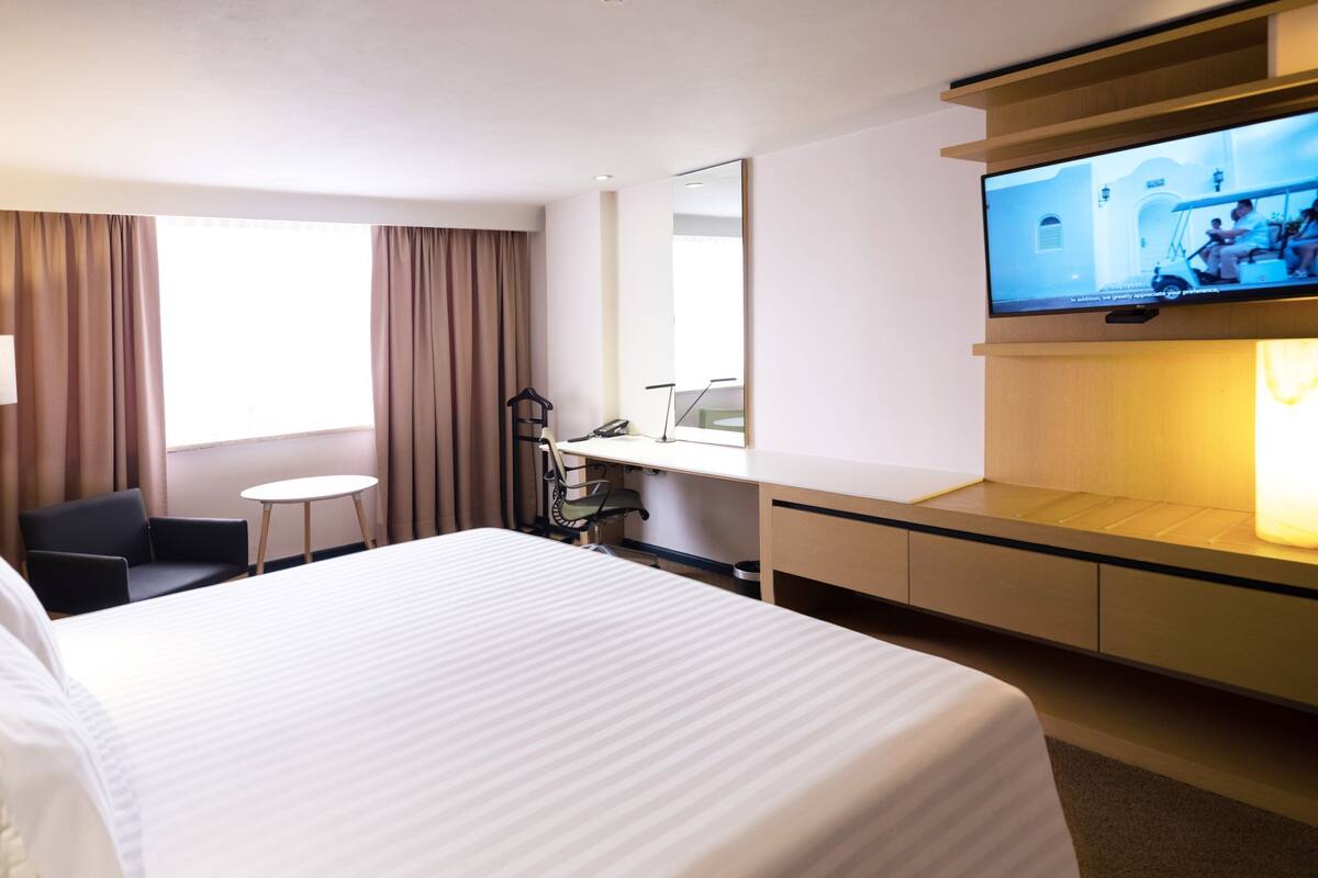 A hotel room with a large bed, a desk, a chair, a small table, a TV, and a window with curtains.