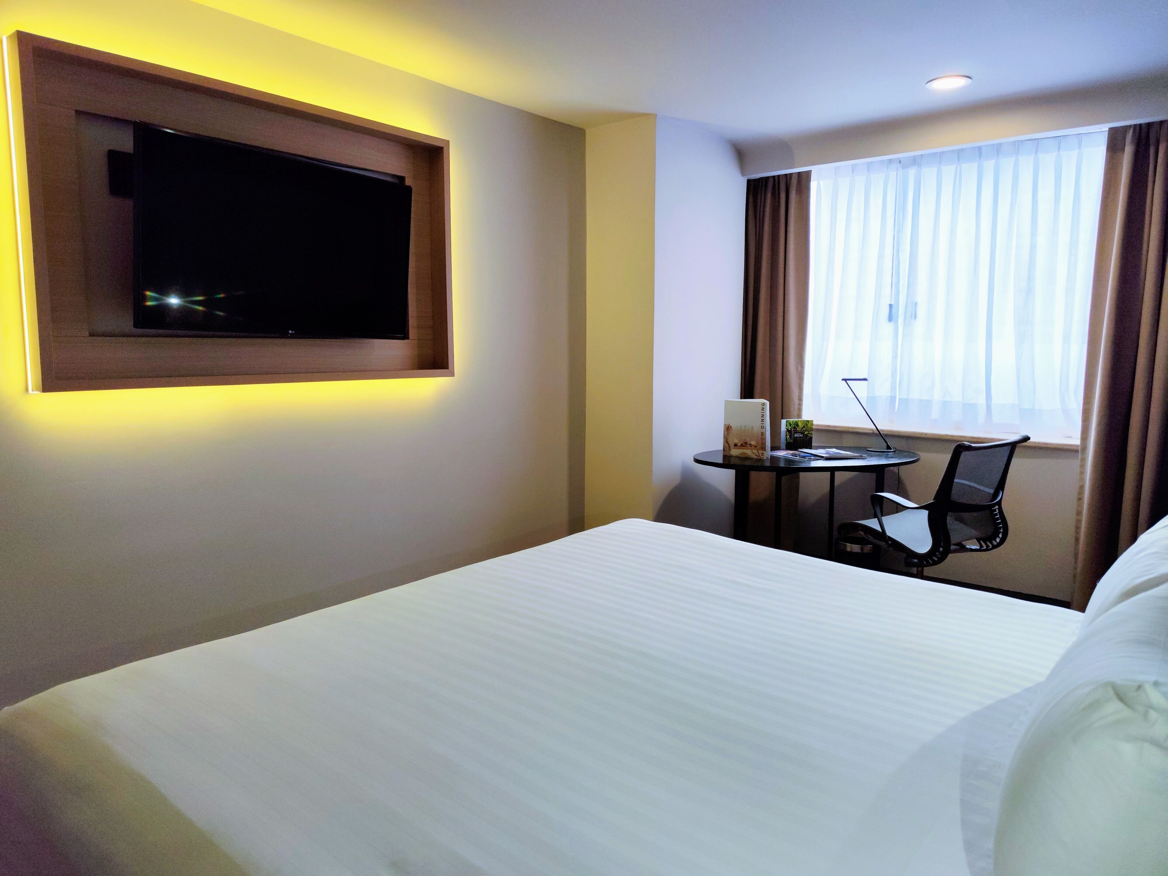 A hotel room with a bed, a TV mounted on the wall, a desk with a chair, and a window with curtains.