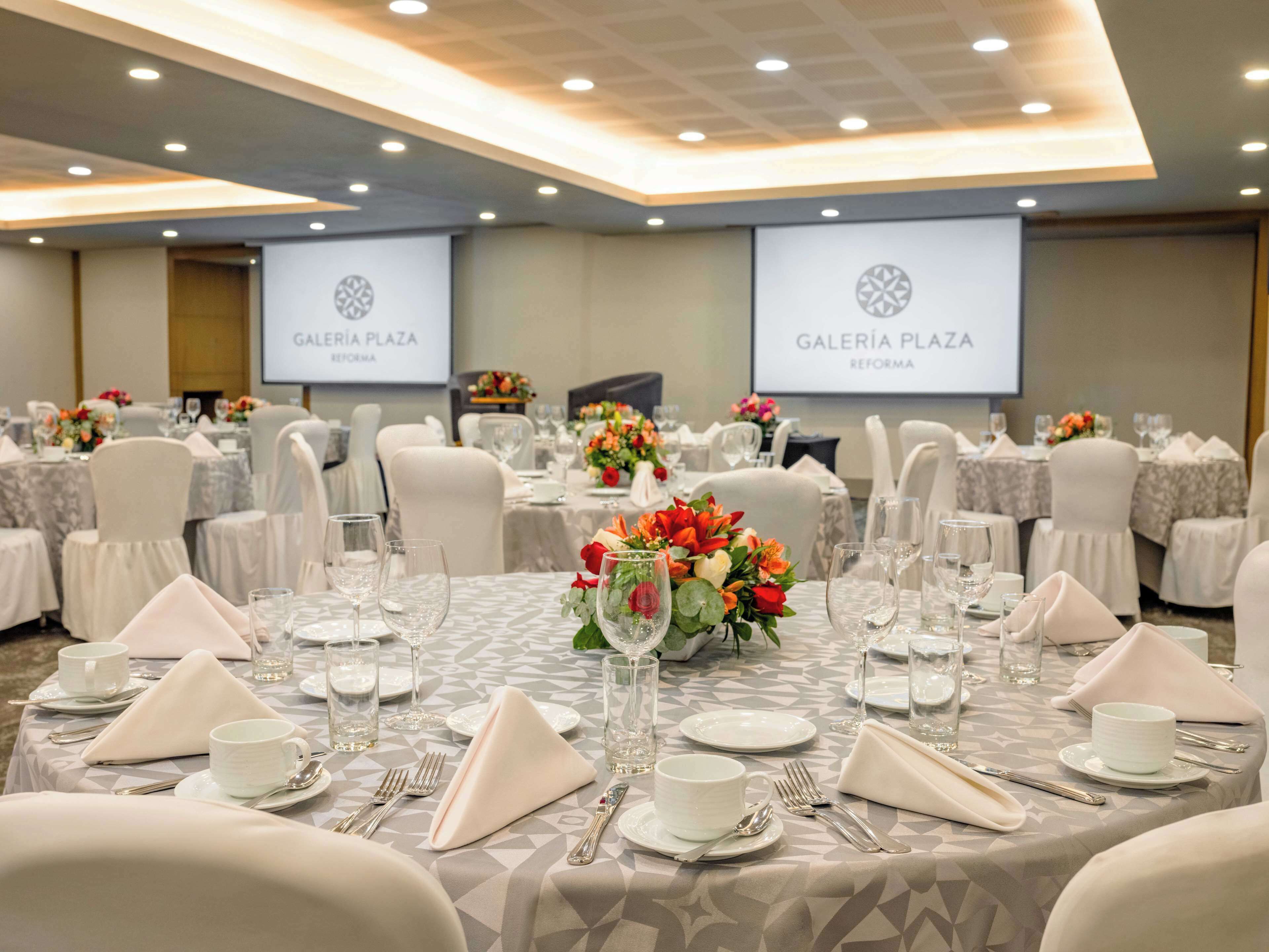 Une salle de banquet où les tables sont dressées pour un événement formel, avec des nappes à motifs géométriques, des chaises blanches et des centres de table floraux.