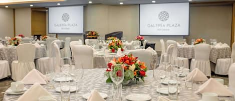 Une salle de banquet où les tables sont dressées pour un événement formel, avec des nappes à motifs géométriques, des chaises blanches et des centres de table floraux.