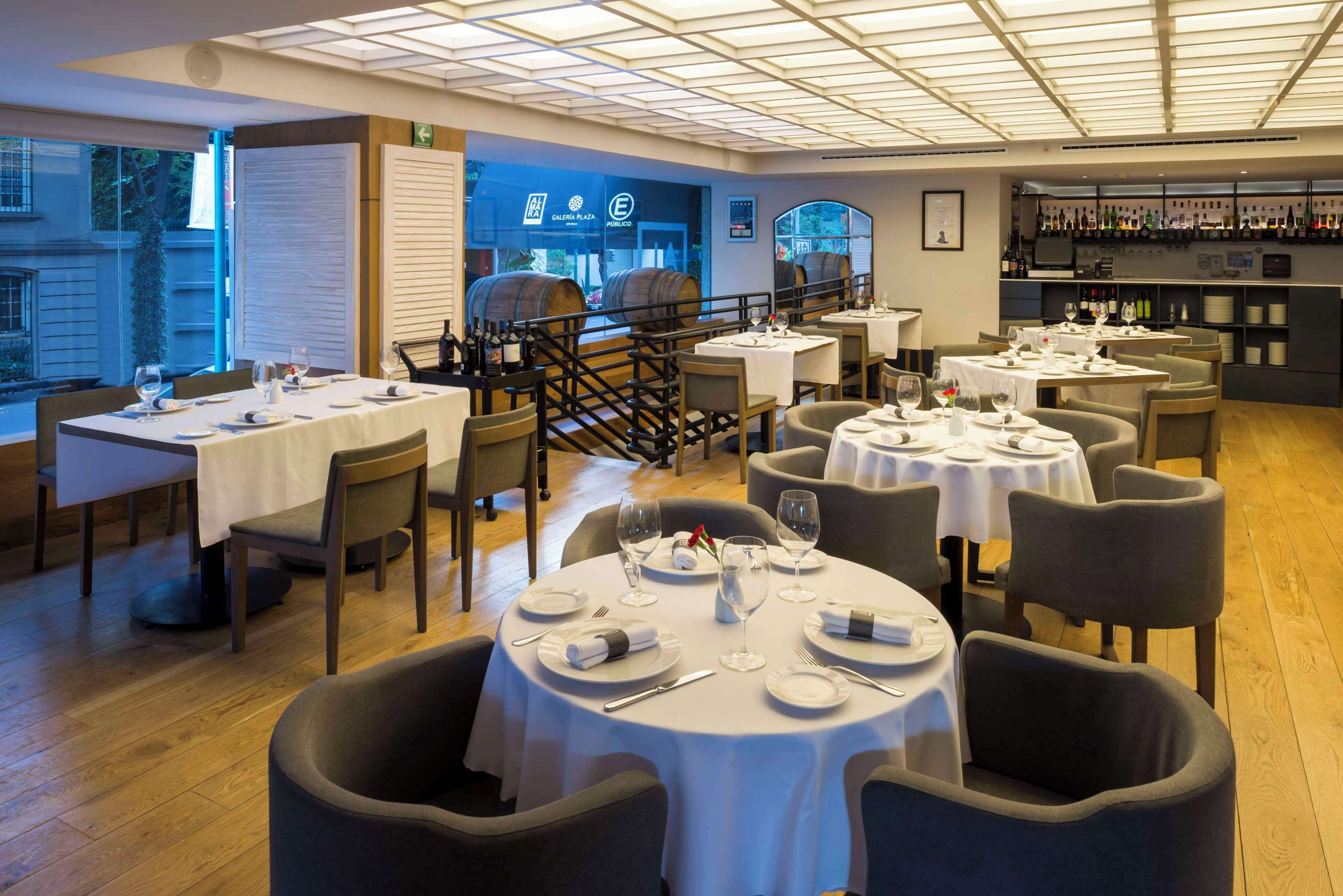 En restaurant med runde borde dækket til spisning, et vinreol i baggrunden og udsigt til gaden udenfor.