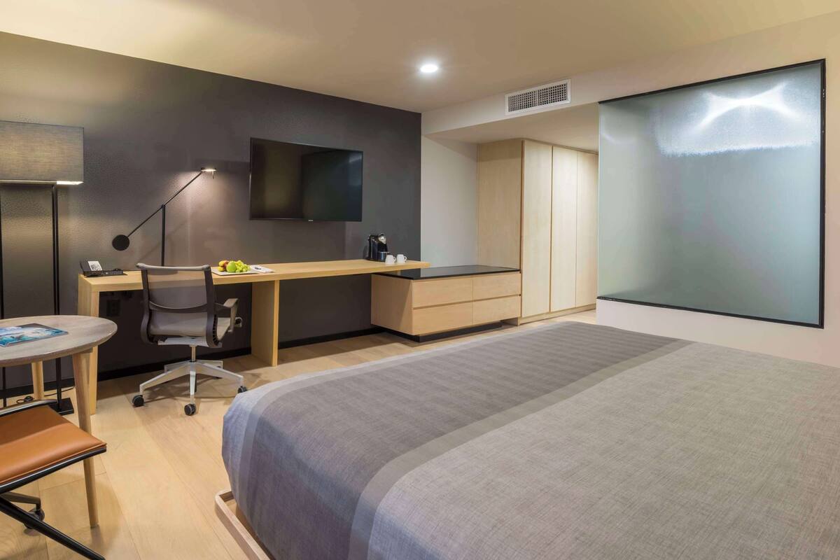 A hotel room with a bed, a desk with a chair, a small table, a TV, and a wardrobe.