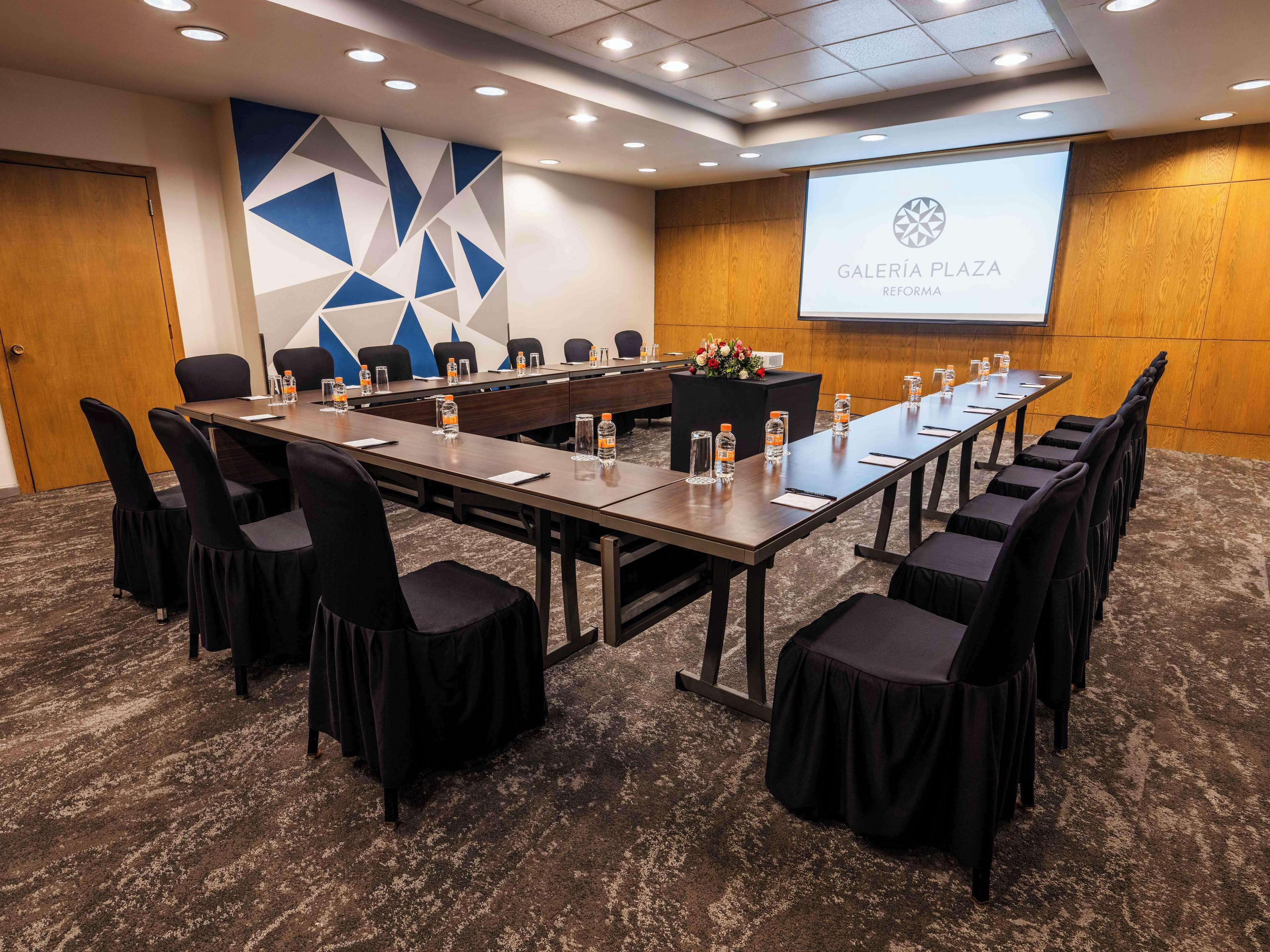 Un salón de conferencias con una larga mesa, sillas, botellas de agua y una pantalla proyectora que muestra 