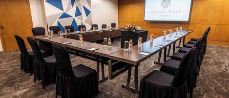 Un salón de conferencias con una larga mesa, sillas, botellas de agua y una pantalla proyectora que muestra