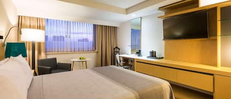 A hotel room with a large bed, a desk with a flat-screen TV, a small table with a lamp, and a view of the city through the window.