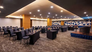 Uma sala de conferências com fileiras de mesas e cadeiras, um palco com fundo azul e um design geométrico na parede.