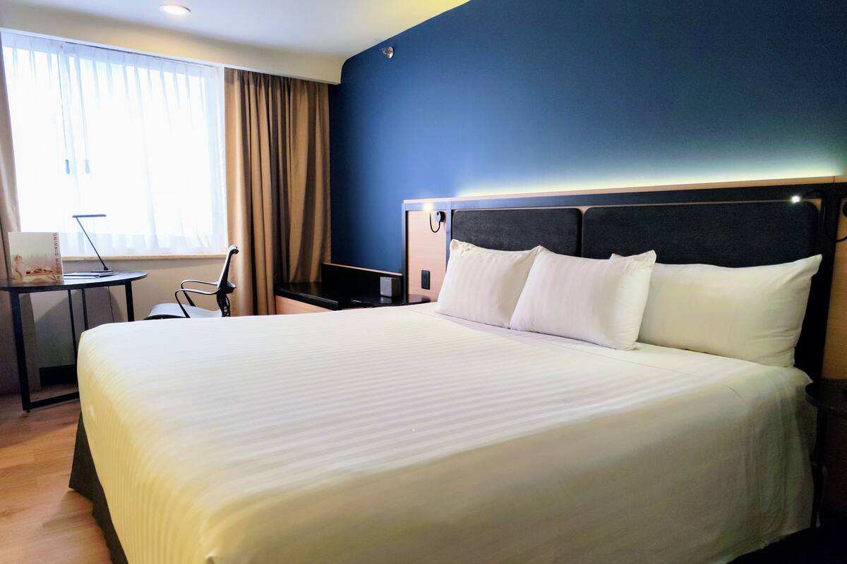 A hotel room with a large bed, a desk, a chair, and a window with curtains.