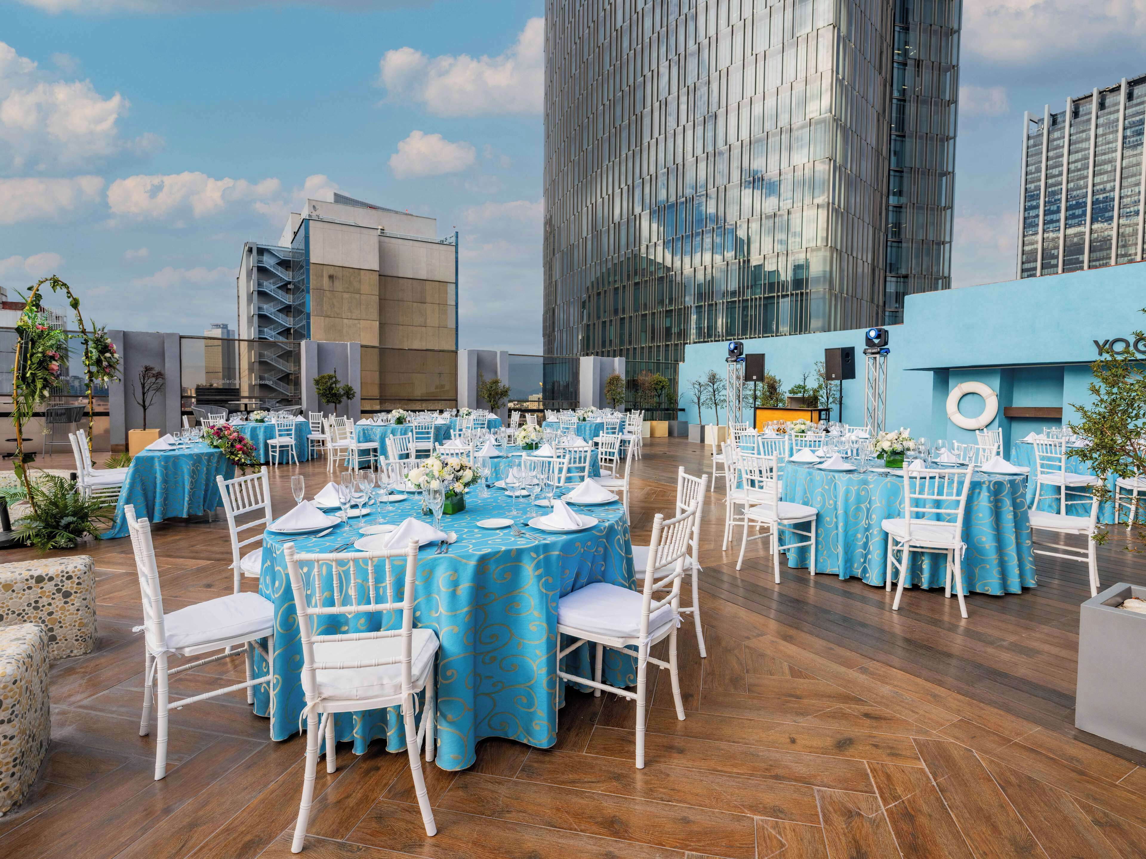 Un área de comedor al aire libre con mesas redondas, sillas blancas y manteles azules.