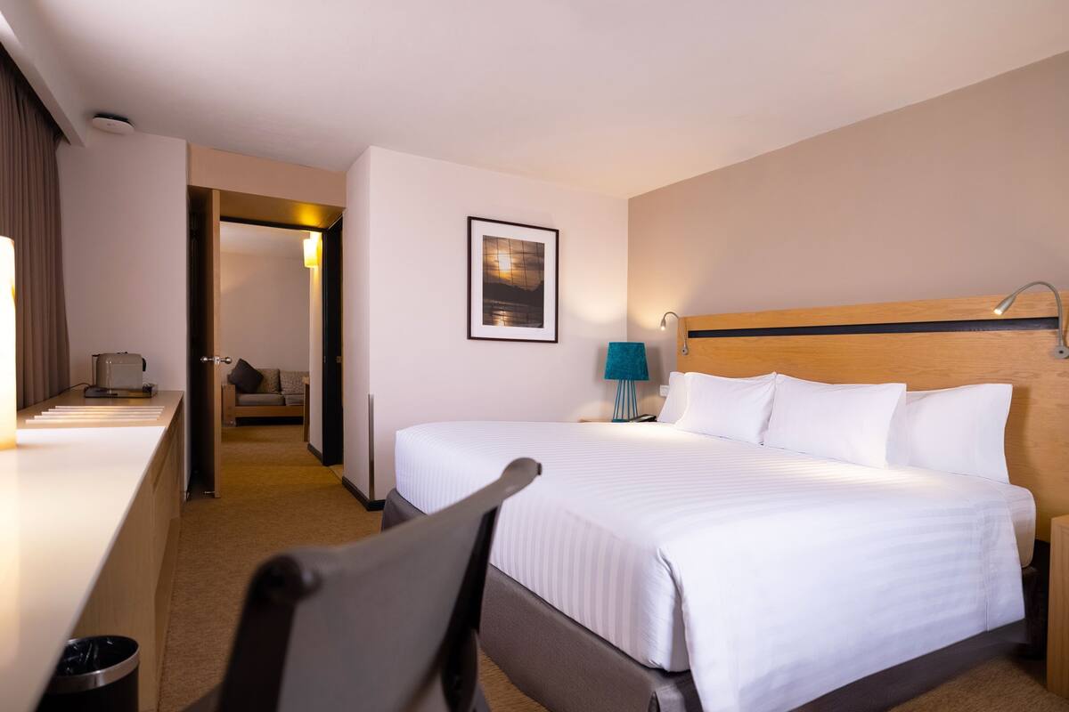 A hotel room with a large bed, a desk with a lamp, a picture on the wall, and a view of the hallway.