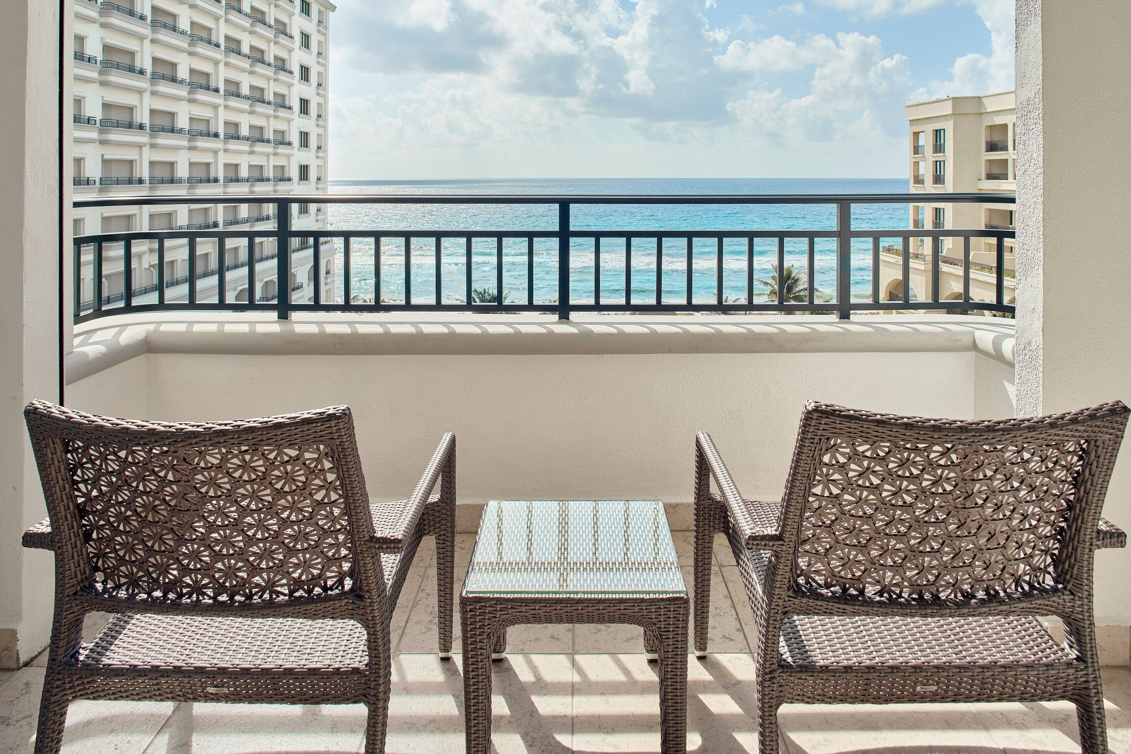 Een balkon met rieten stoelen en een tafeltje, met uitzicht op een strand en de oceaan.
