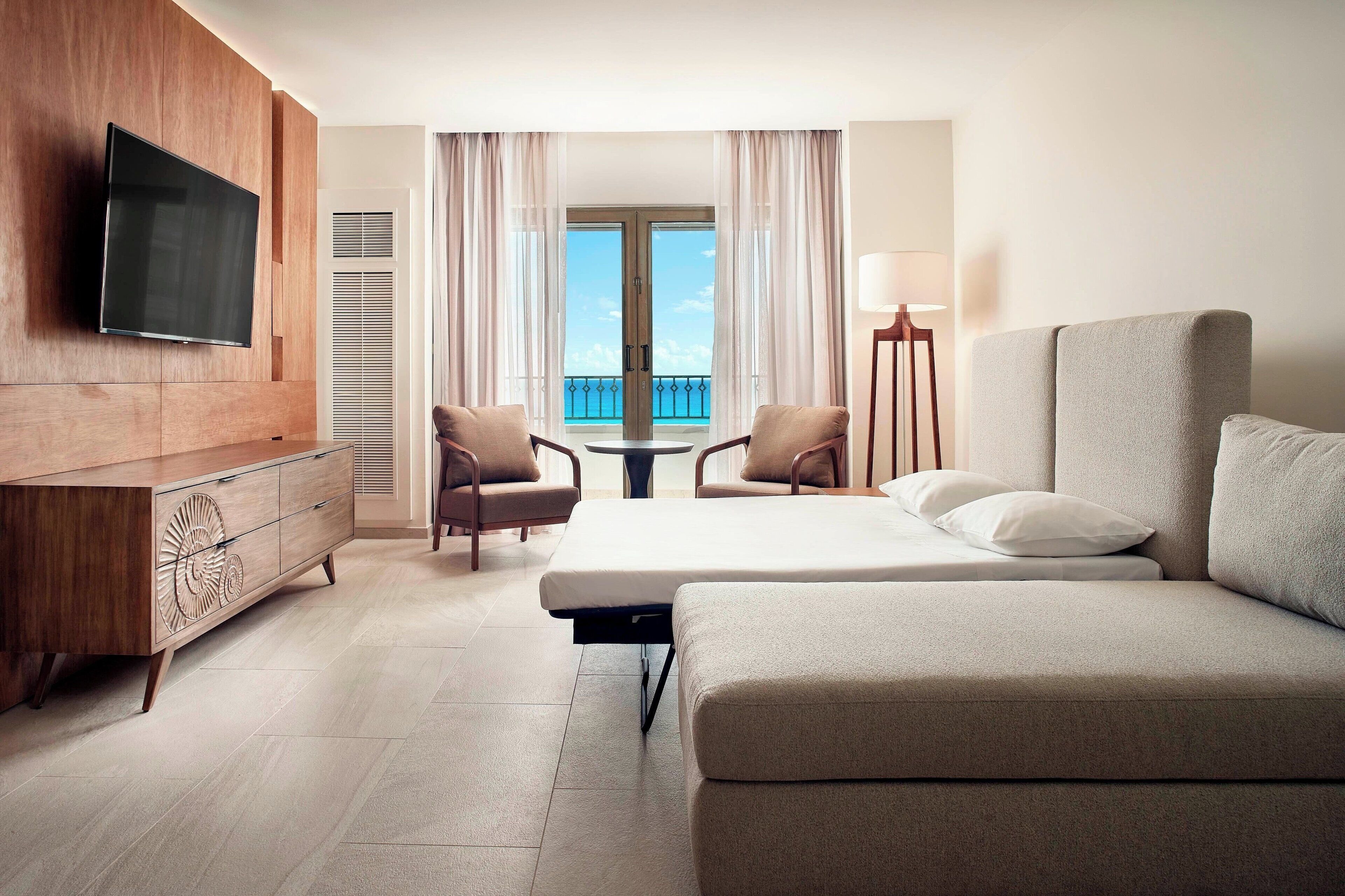 A hotel room with a bed, a TV mounted on a wooden cabinet, two chairs, a small table, and a balcony with a view.