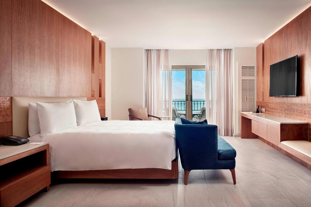 A modern hotel room with a large bed, a seating area with a chair, a TV mounted on the wall, and a balcony with curtains.